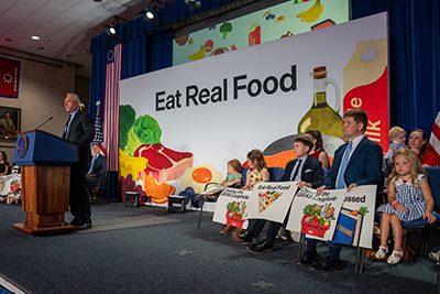 Health Secretary Robert F. Kennedy Jr. stands at a podium in front of a backdrop that says "Eat Real Food" and has images of various meats, vegetables and other foods on it. Behind him and to one side are children holding signs.